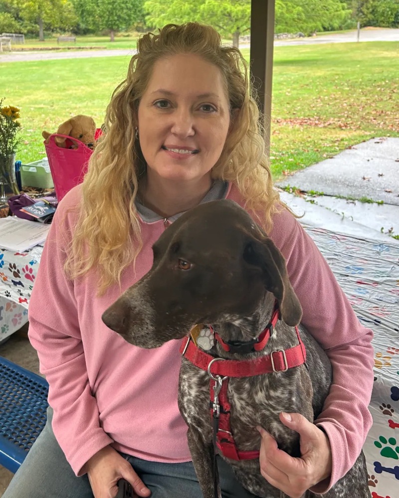Nancy Arnold sitting under a park pavilion with her dog Scout, a brown and white dog in a red harness